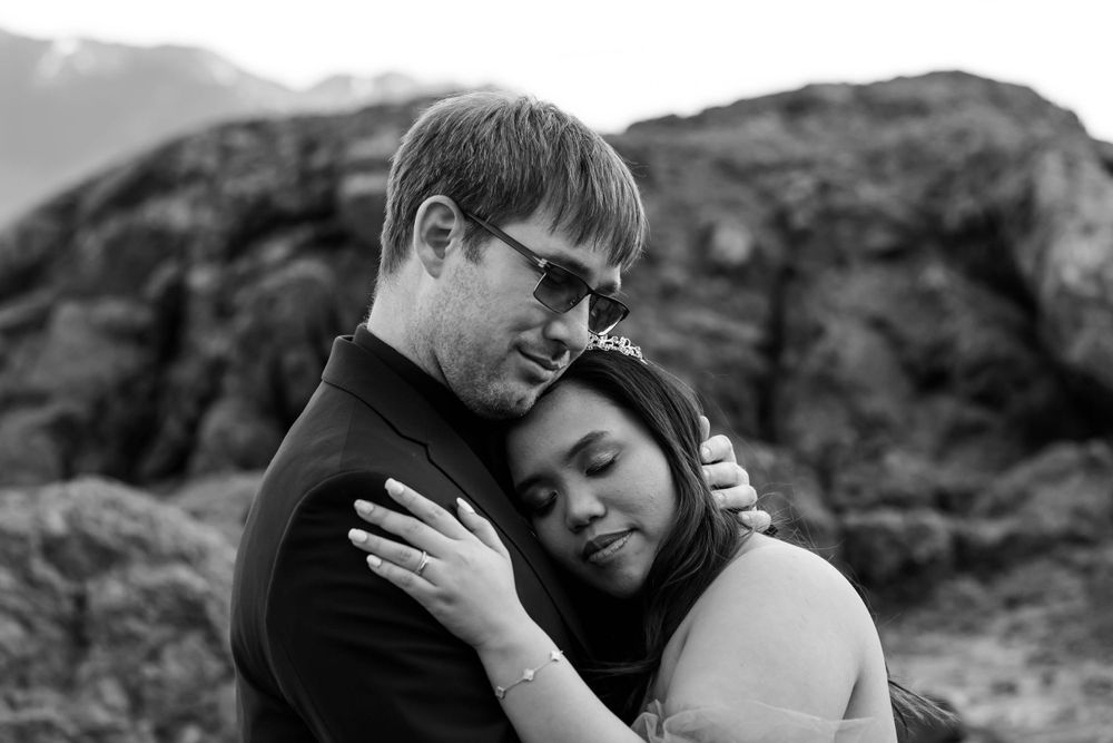 A black and white portrait of a couple sharing a tender embrace against mountain scenery.