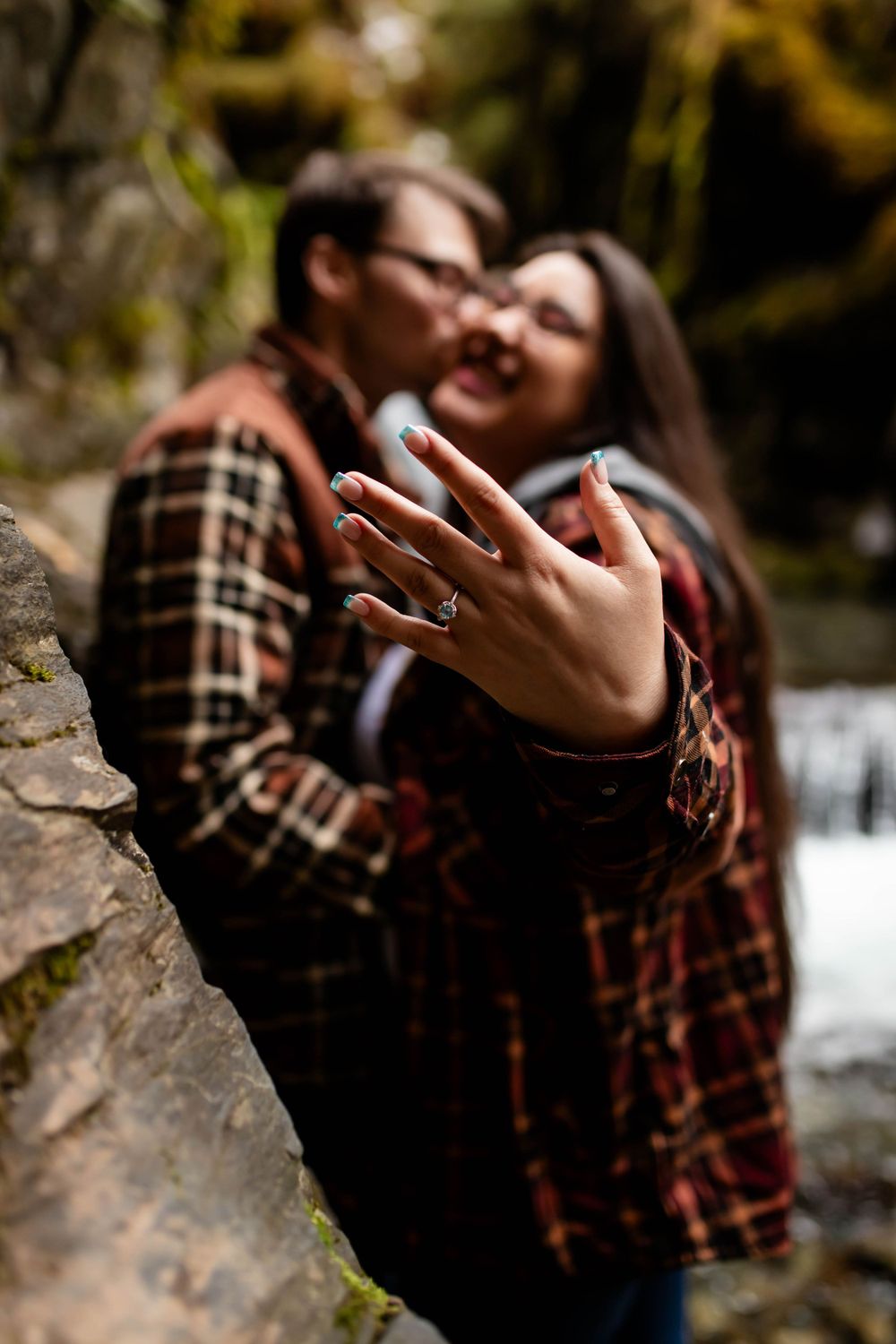 A close-up of an engagement ring while a couple embraces near a waterfall.