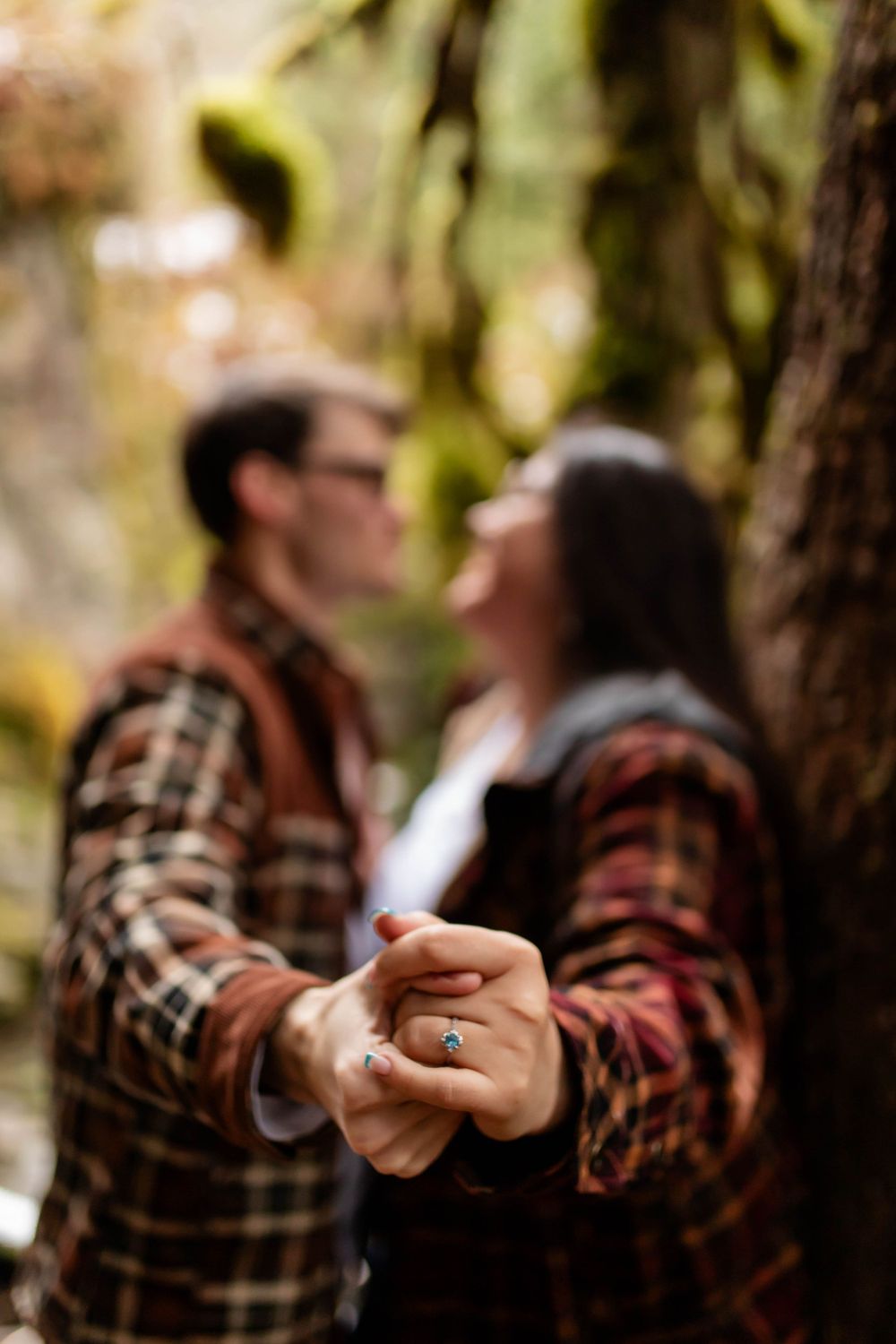 A couple in matching plaid flannel shirts shares an intimate moment in a forest setting.