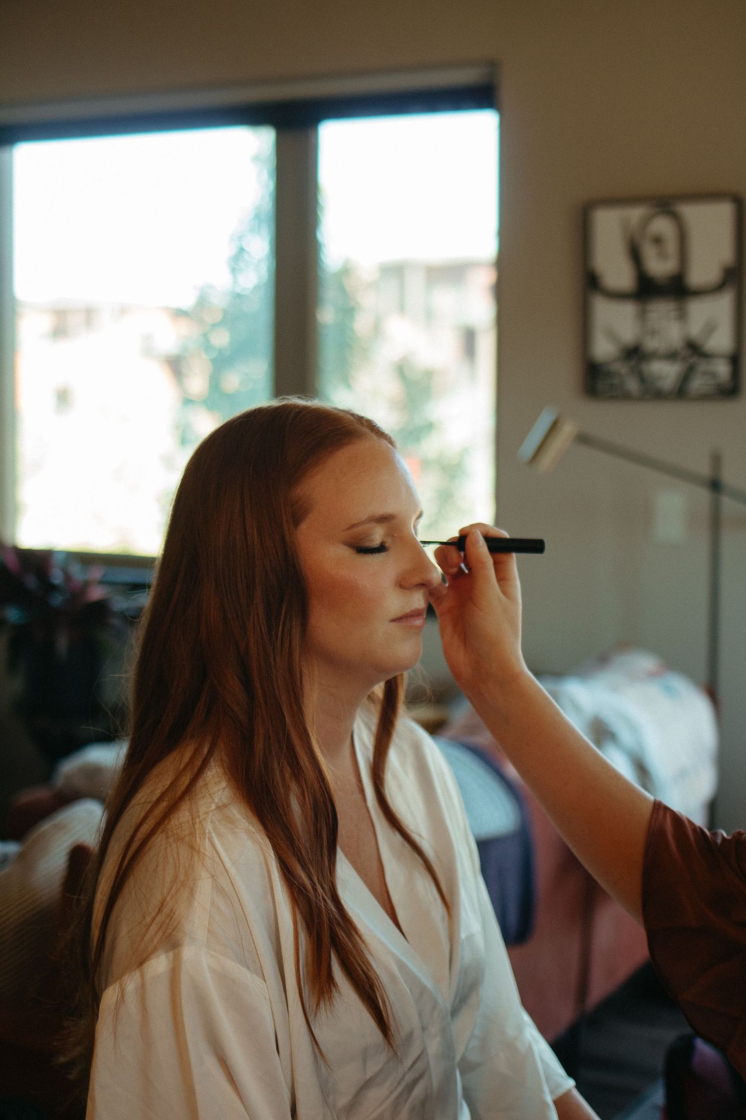 Makeup artist applying cosmetics to client in preparation setting with natural window light.