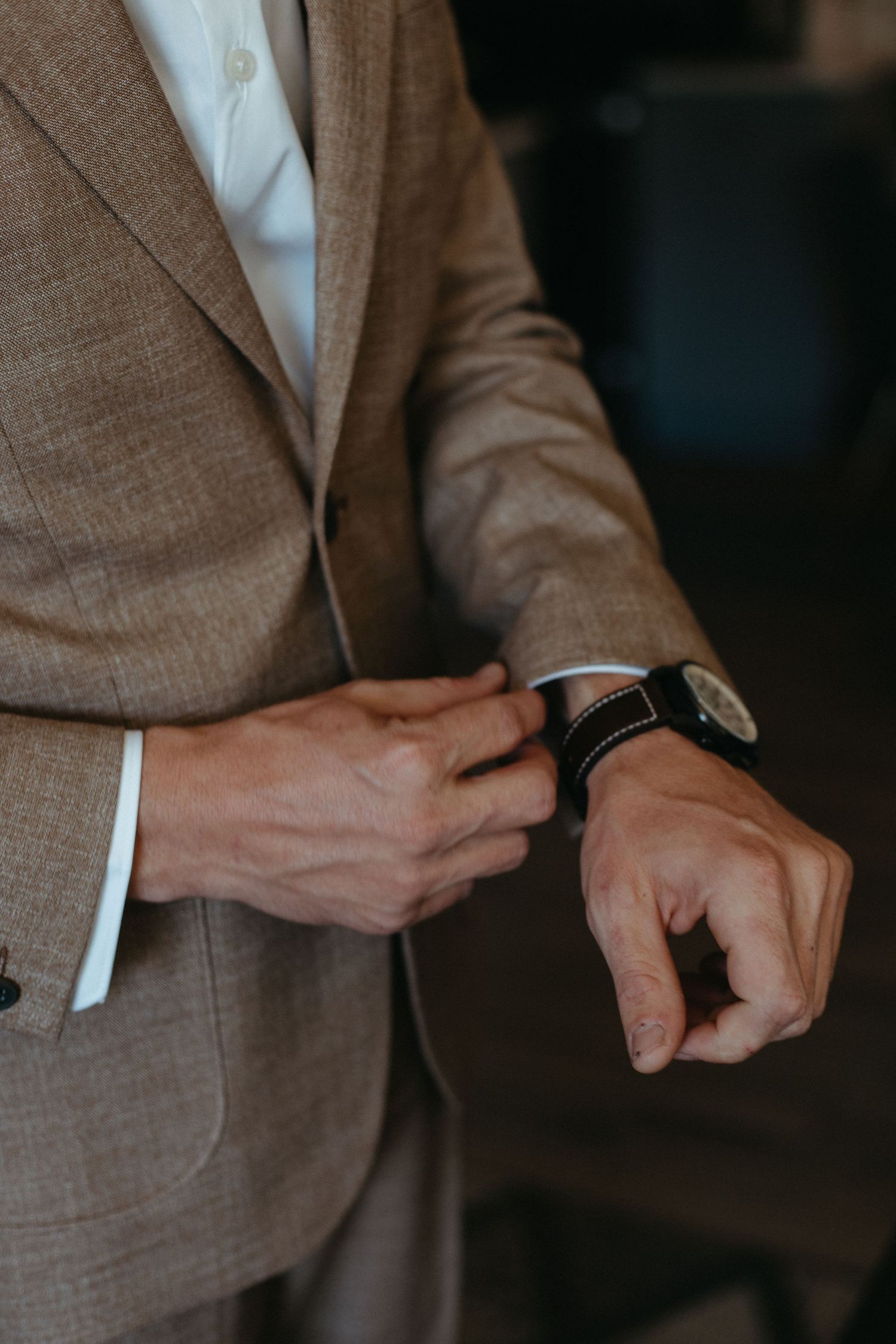 Close-up of hands adjusting a wristwatch while wearing a tan suit jacket.