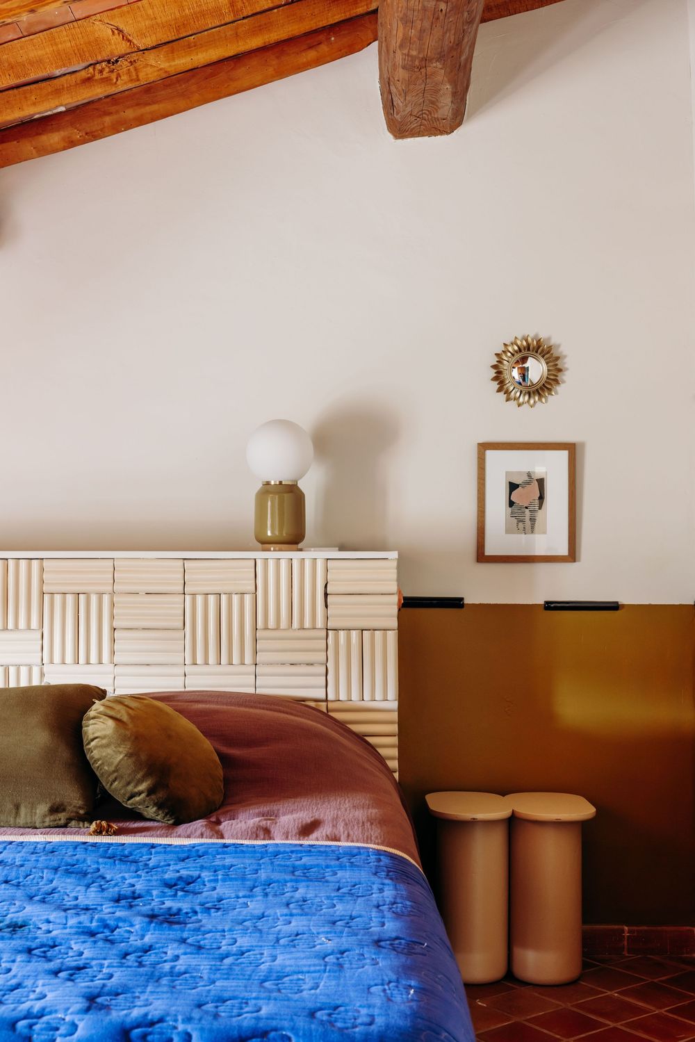 Bohemian bedroom with blue quilt, wooden headboard, rattan dresser, globe lamp, gold mirror, and exposed beam ceiling.