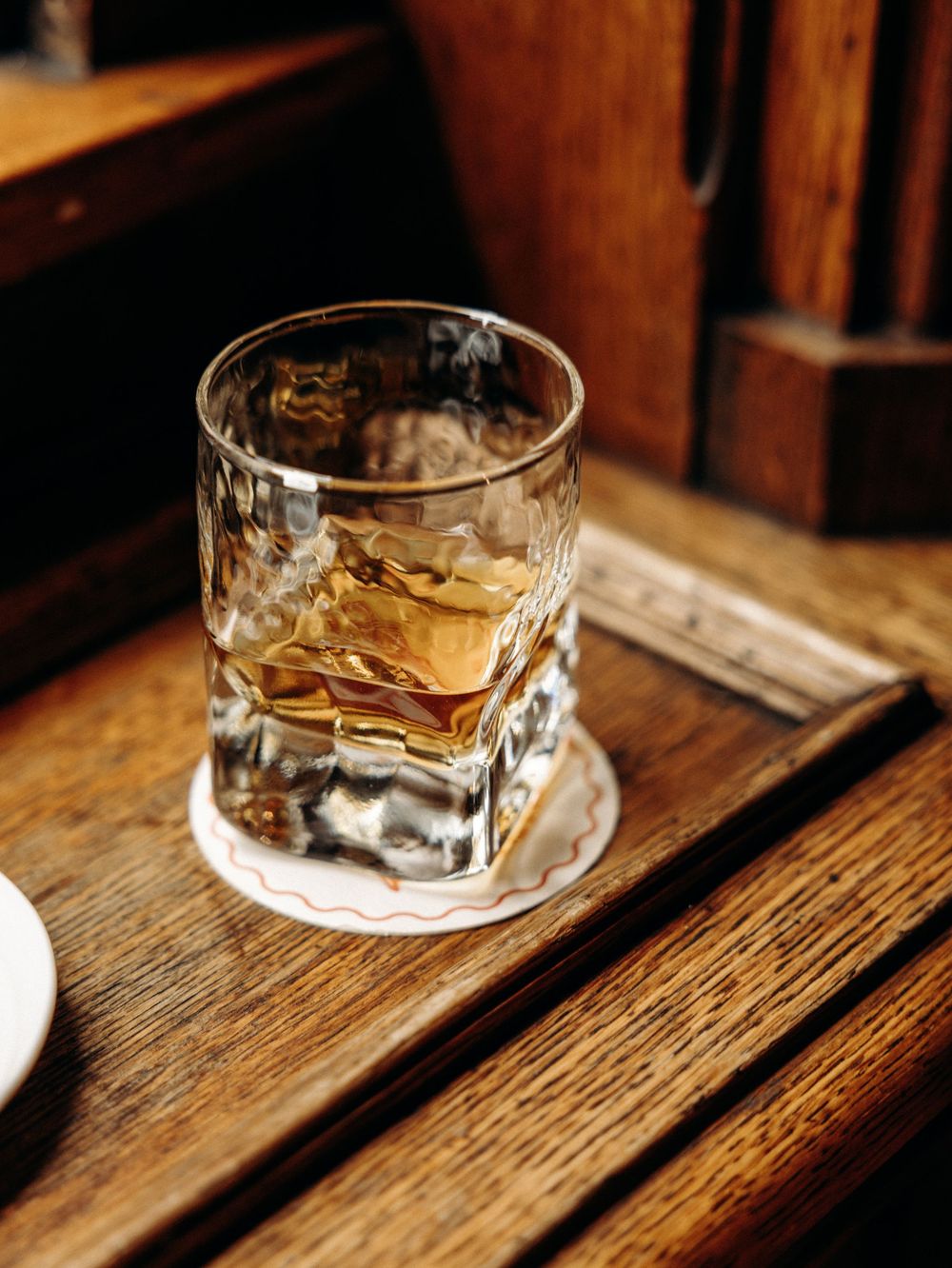 Crystal glass of whiskey with ice on a wooden coaster atop a rustic wood table in a dimly lit bar.