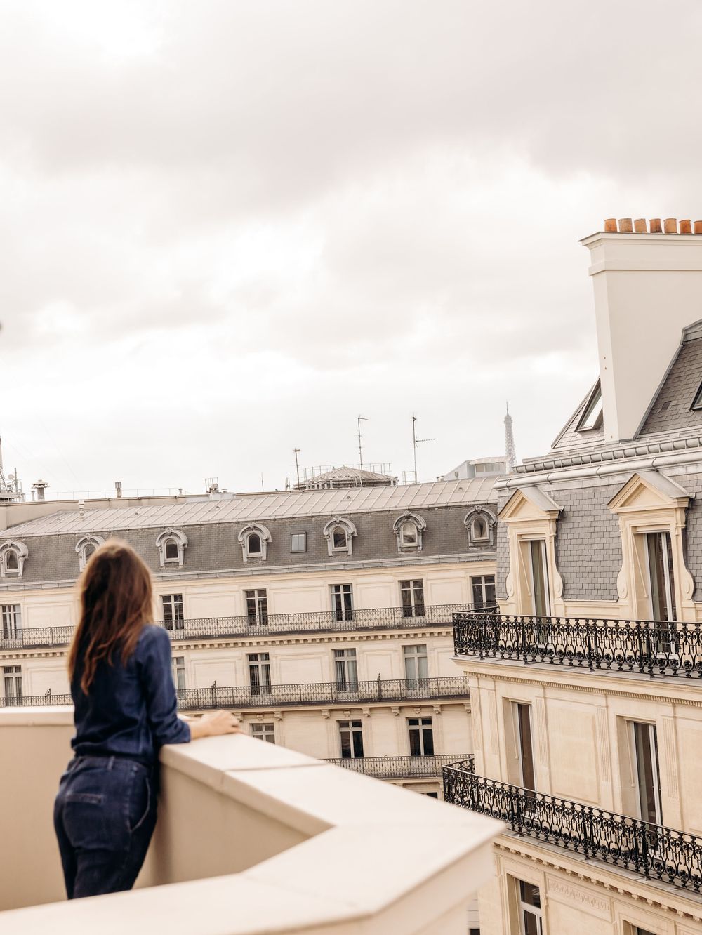 A woman in denim stands on a Parisian rooftop balcony, gazing at classic Haussmann architecture under cloudy skies.