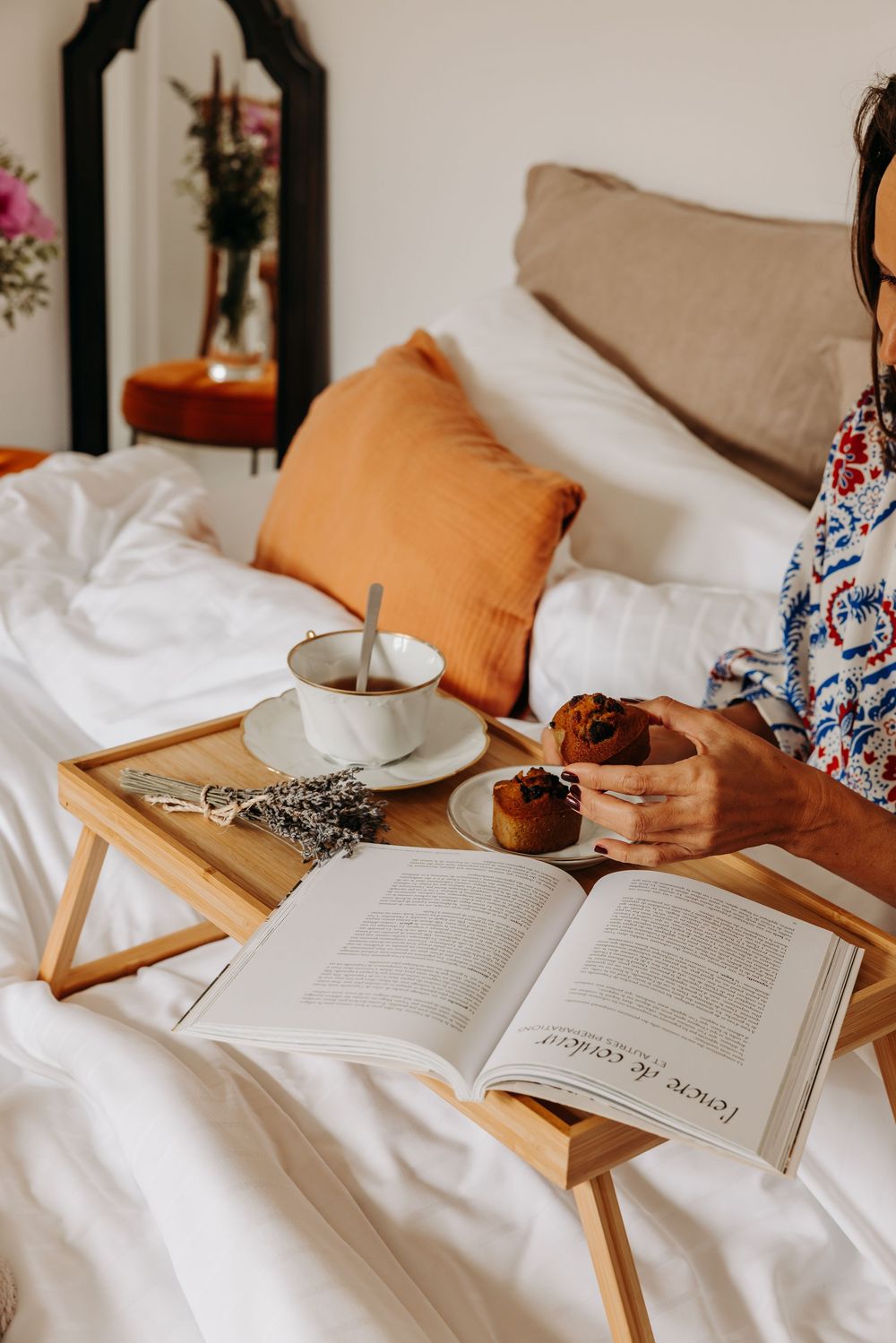Woman enjoying breakfast in bed with coffee, muffin, open book, and lavender on a wooden tray.