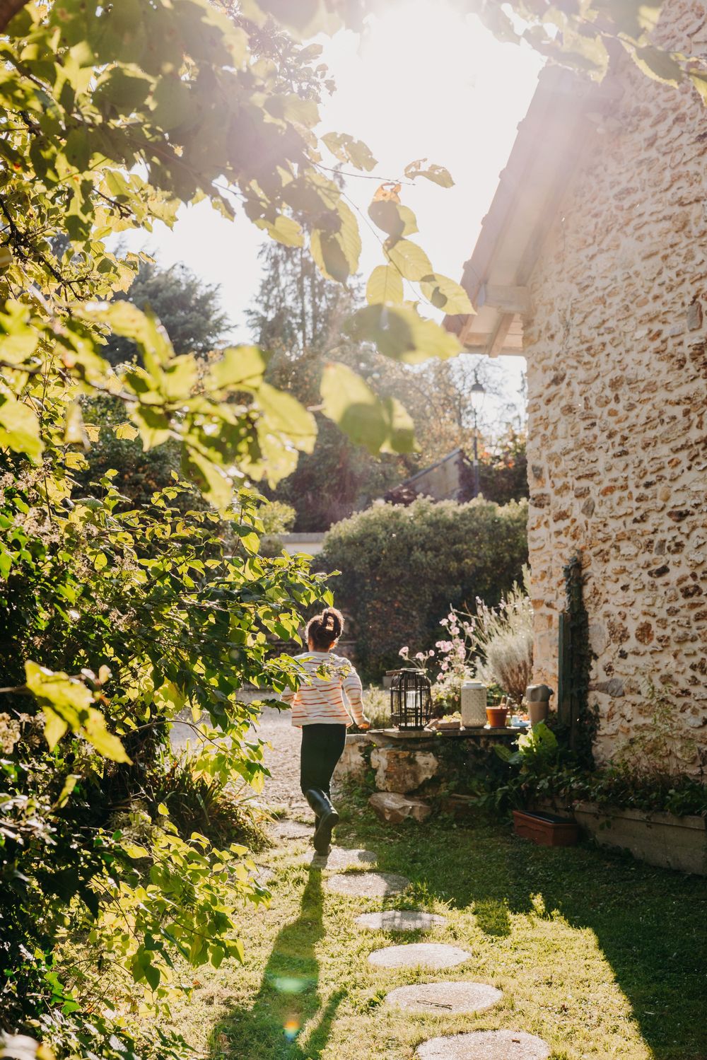Woman walking along a sunlit stone cottage garden path surrounded by lush green foliage on a bright sunny day.