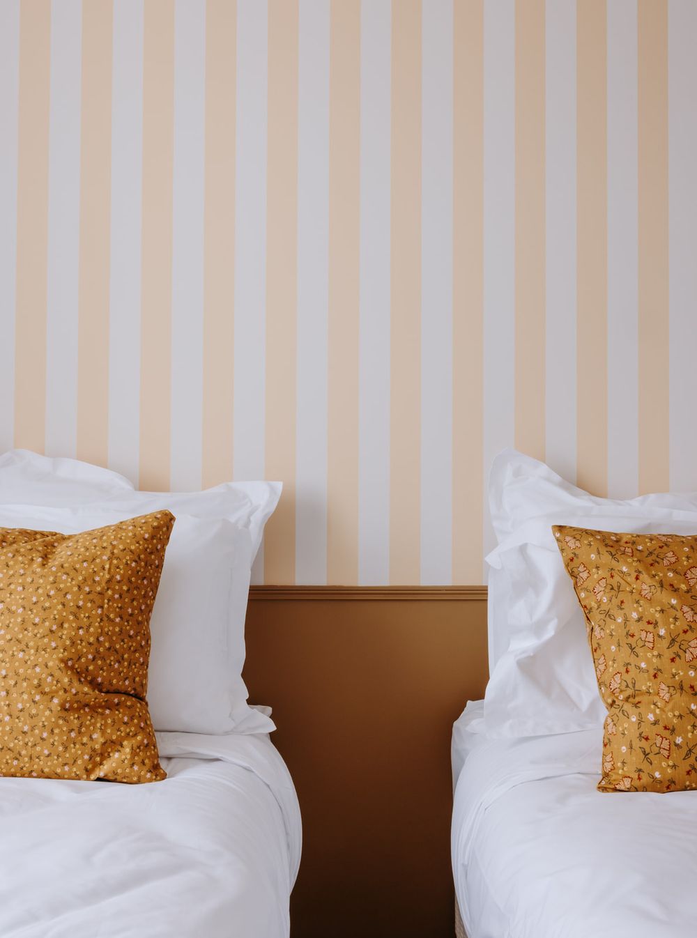 Two twin beds with white linens and gold floral pillows against a striped peach and white wallpaper.