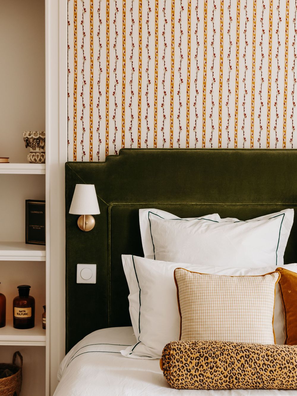 Cozy bedroom with green velvet headboard, leopard print pillow, and chain-pattern wallpaper with brass sconce.