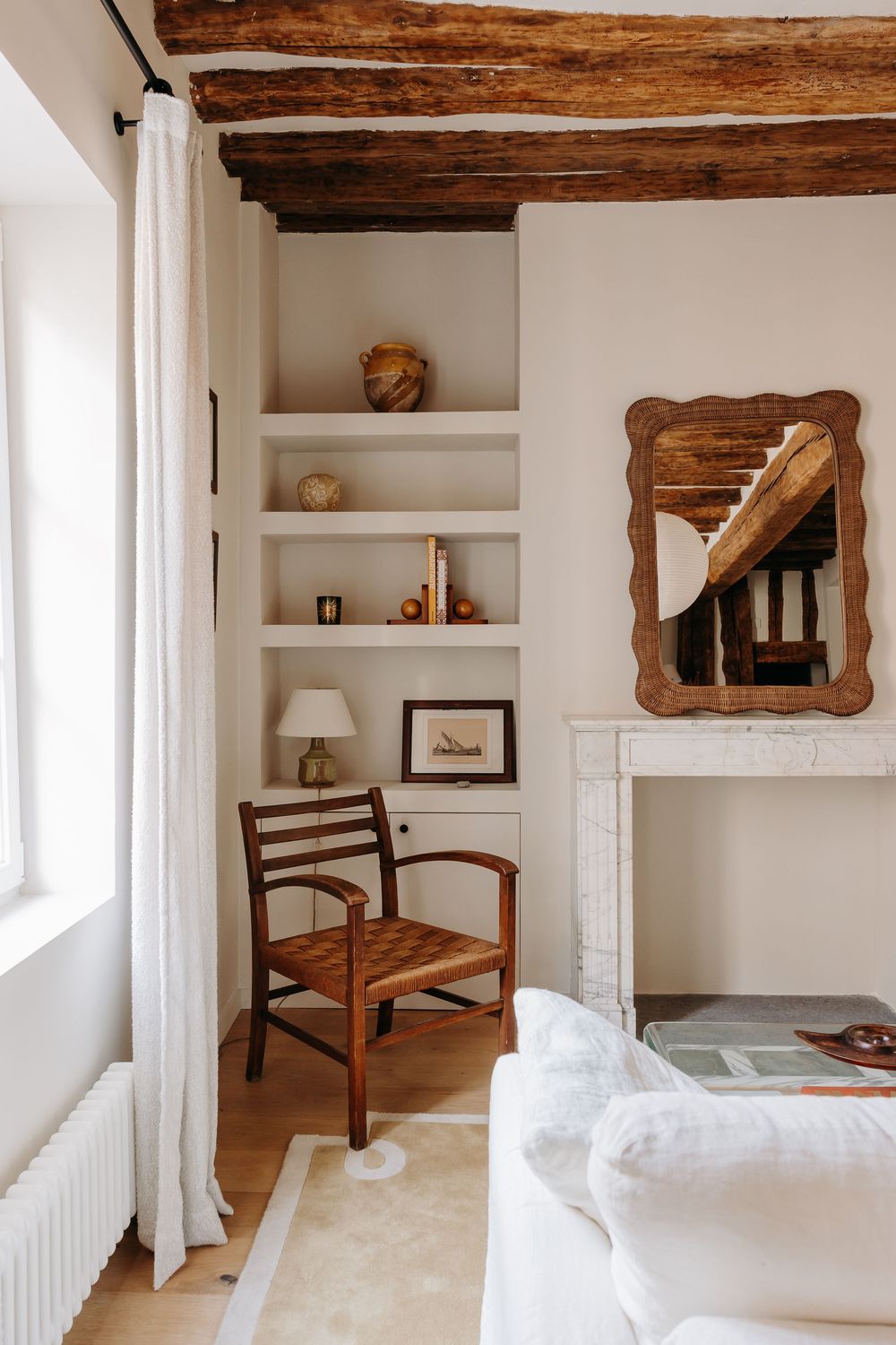 Rustic French living room with exposed wood beams, built-in shelves, antique chair, and white fireplace mantel.