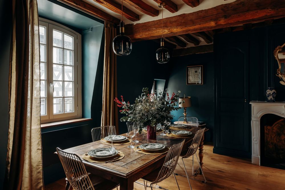 Rustic dining room with dark teal walls, wooden beam ceiling, farmhouse table, mixed chairs, and floral centerpiece near a window.