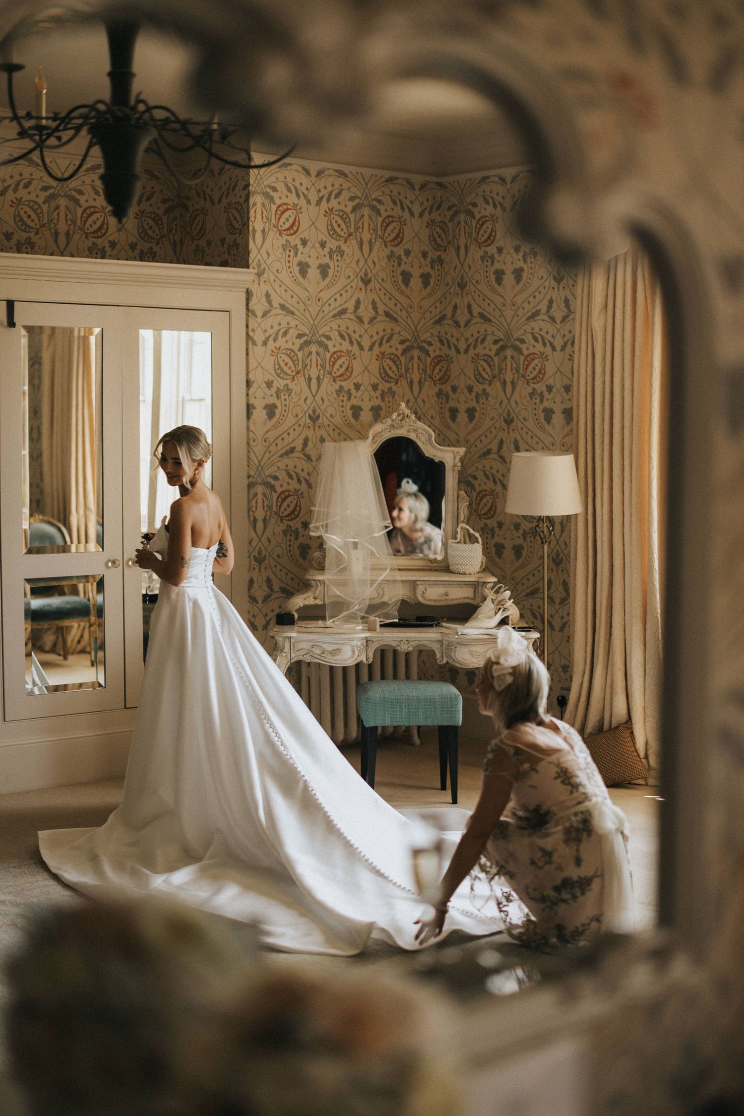 A bride in a flowing white wedding gown gets ready in an elegant room at Hodsock Priory with vintage floral wallpaper.
