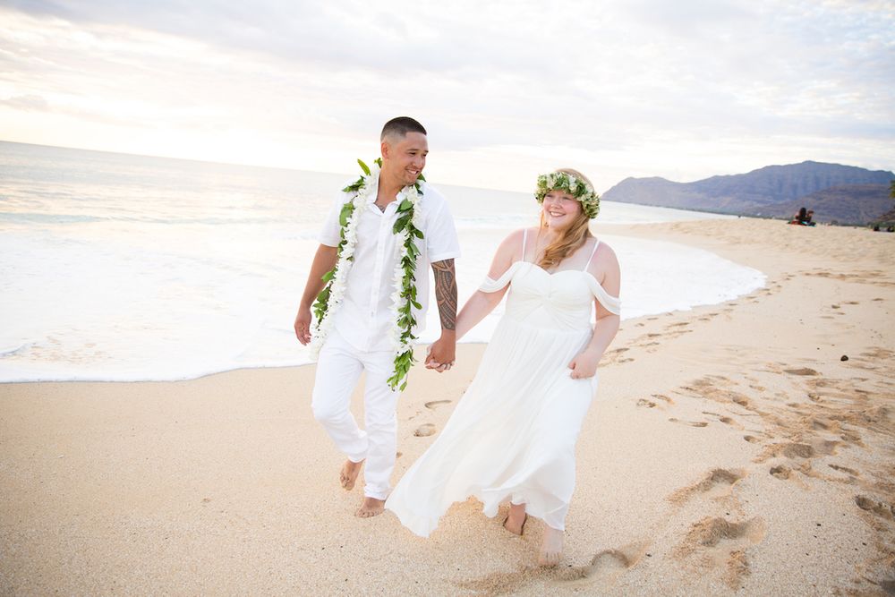 A couple in white wedding attire walks hand in hand along a sandy beach with mountains in the background of Māʻili Beach Park.
