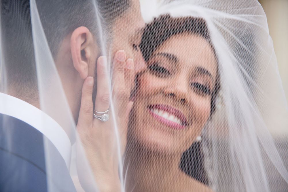 A bride in a white veil smiles joyfully while sharing an intimate moment with her groom on their wedding day.