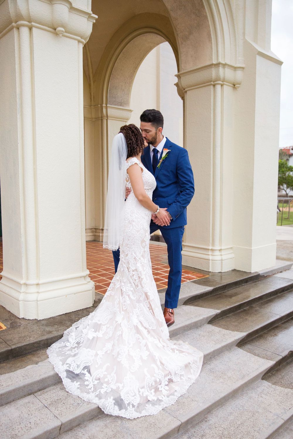 A bride in a lace gown with long train and groom in navy suit embrace on elegant white stone steps with arches.