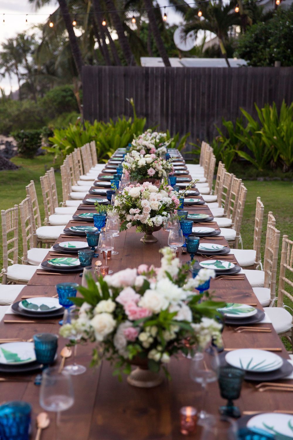 Elegant outdoor wedding reception table with blue glassware and white floral centerpieces set against tropical landscaping.