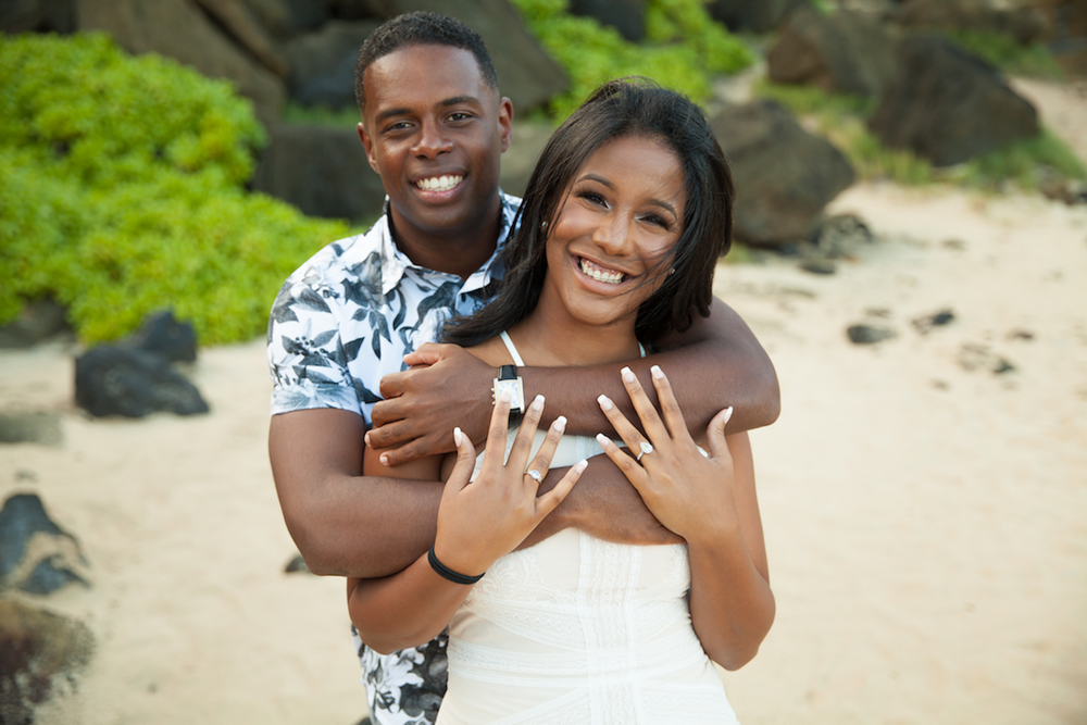 A happy couple poses on a beach, with the bride in a white dress showing off her ring while being embraced from behind.