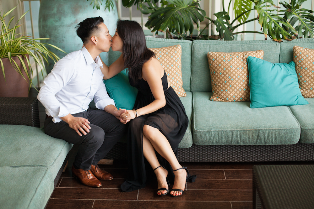 A romantic couple shares an intimate moment on a teal couch with decorative gold pillows in a modern living space.