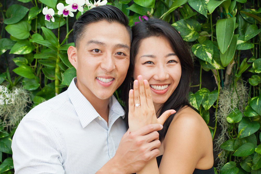 Happy couple showing off engagement ring while posing against green leafy background outdoors.