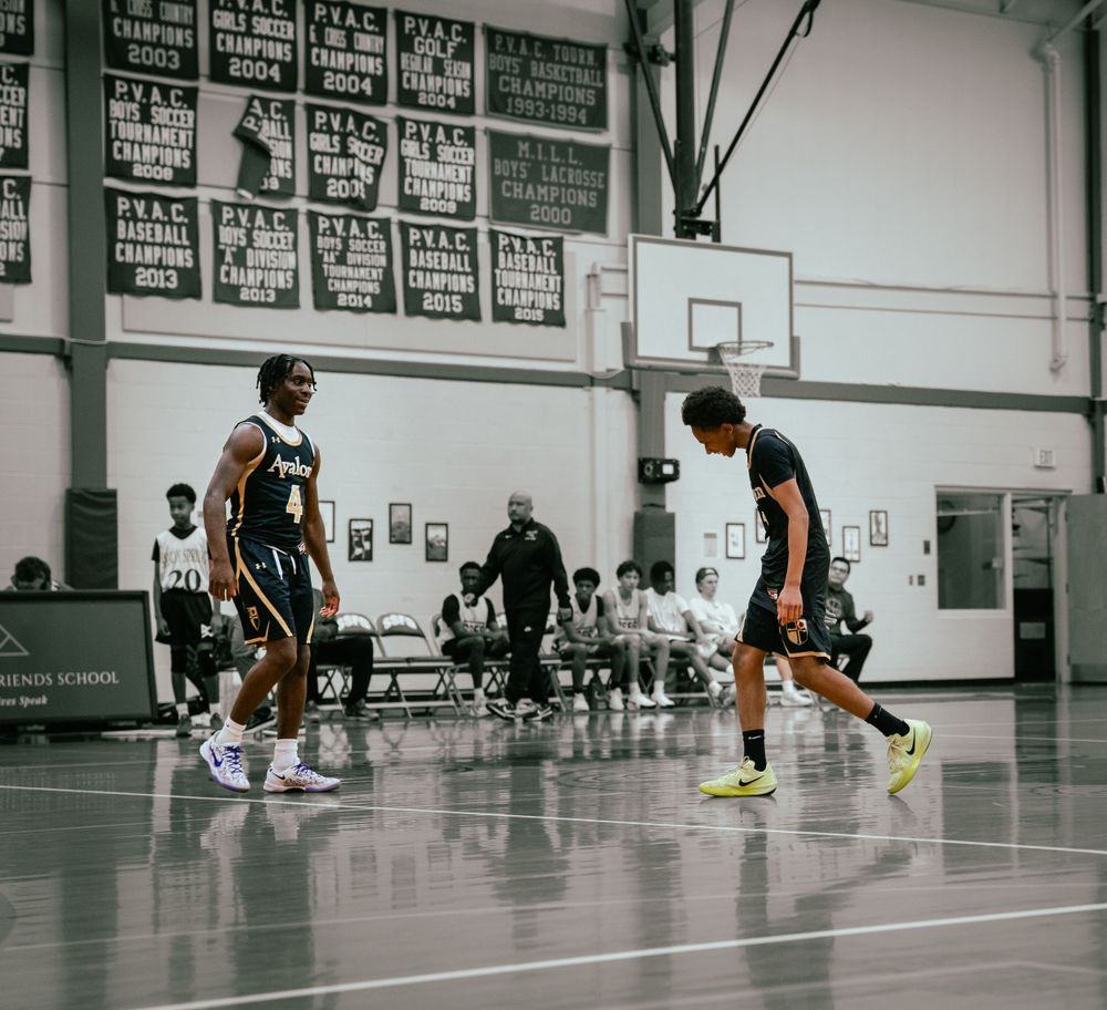 Two basketball players face off on indoor court with championship banners on wall.