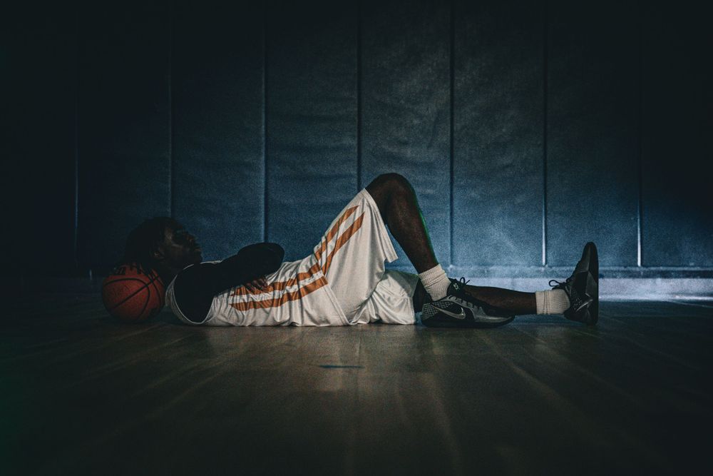 Basketball player rests on gym floor beside a ball against dark padded wall.