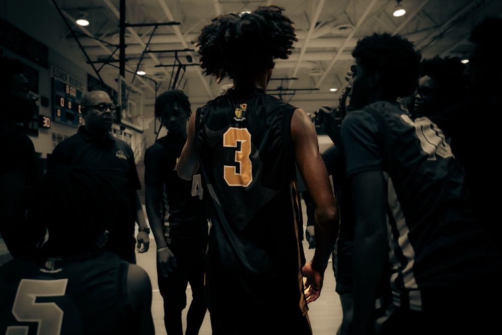 Basketball player wearing number 3 gold jersey walks with teammates through a dimly lit arena.
