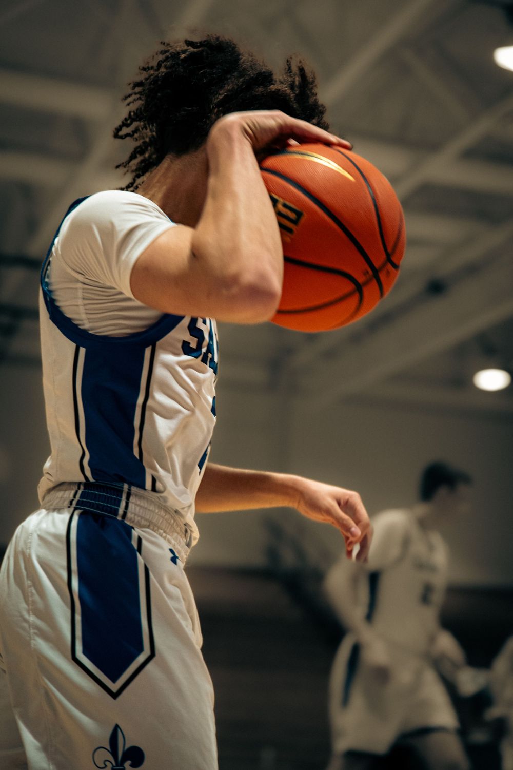Basketball player in white blue jersey holds orange ball near face on indoor court.