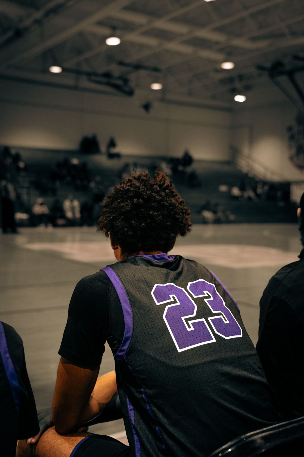 Basketball player wearing number 23 purple jersey sits on the bench watching an indoor game.