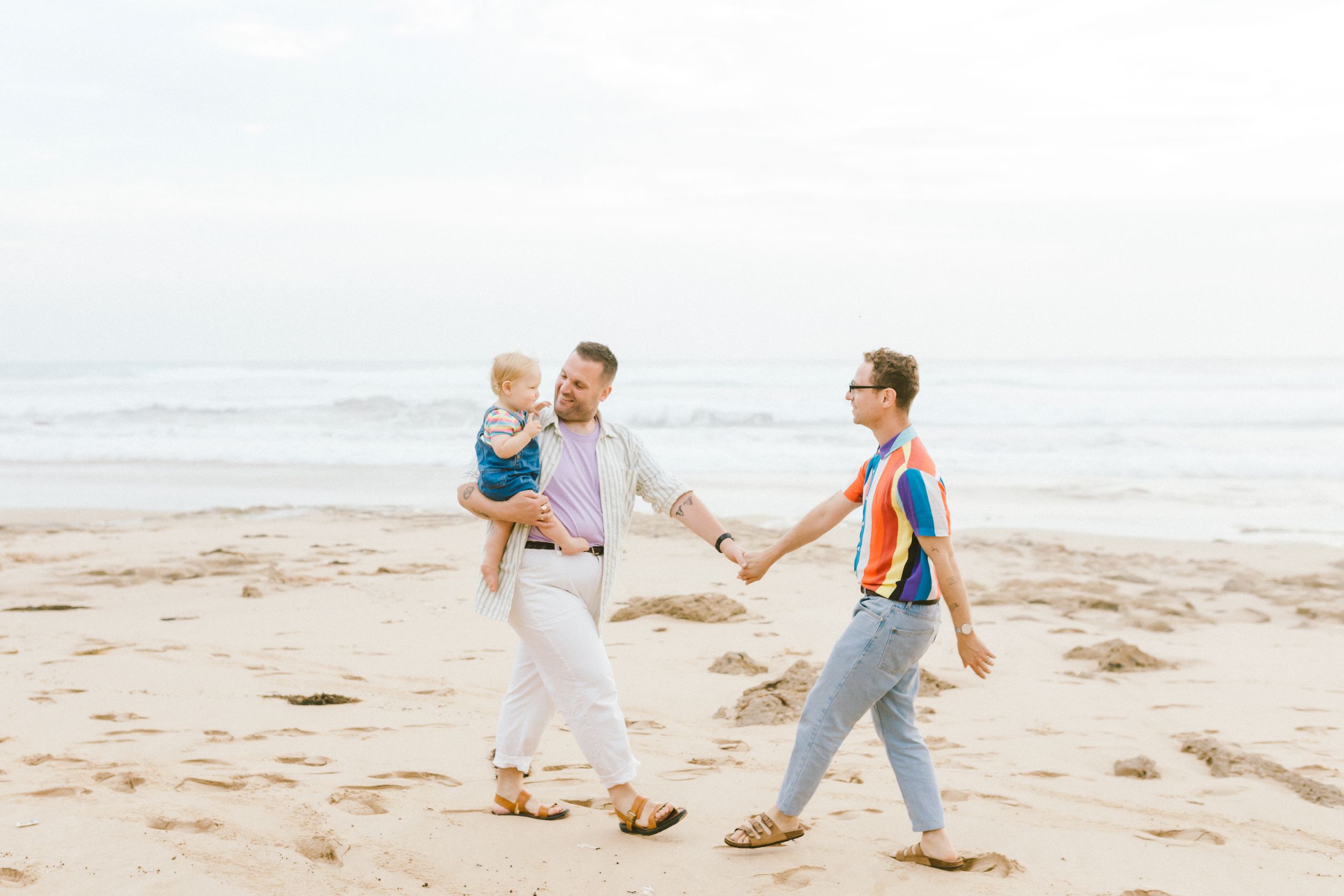 Couple walking hand in hand along a sunny beach with ocean waves in the background.