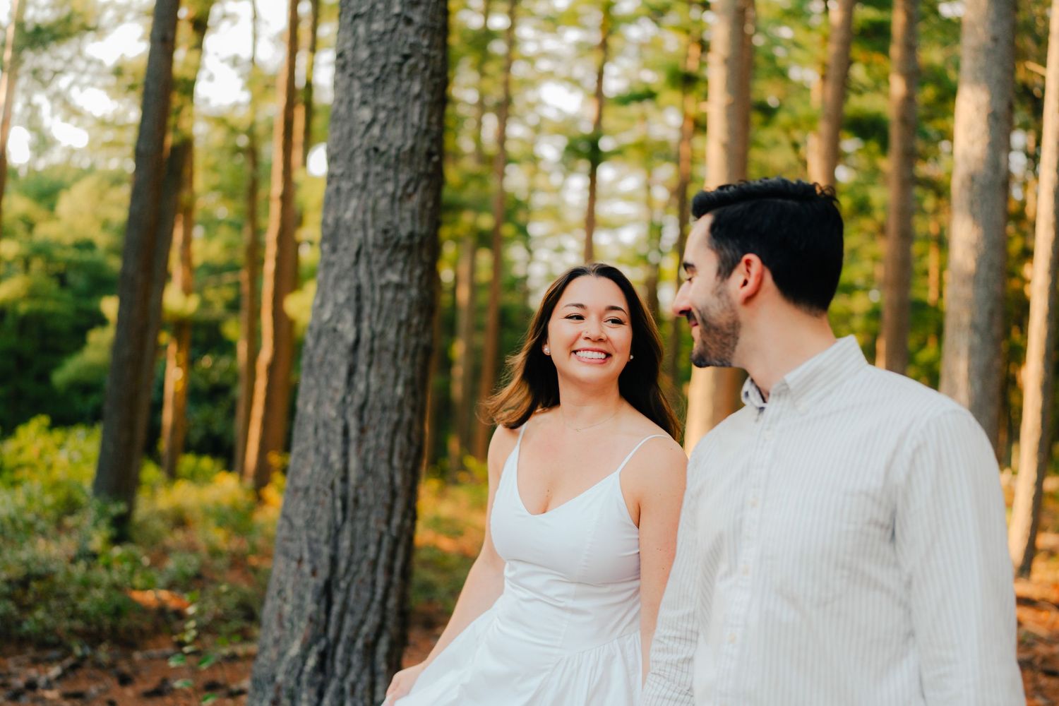 A couple in wedding attire walks through a sunlit forest with tall pine trees during golden hour.