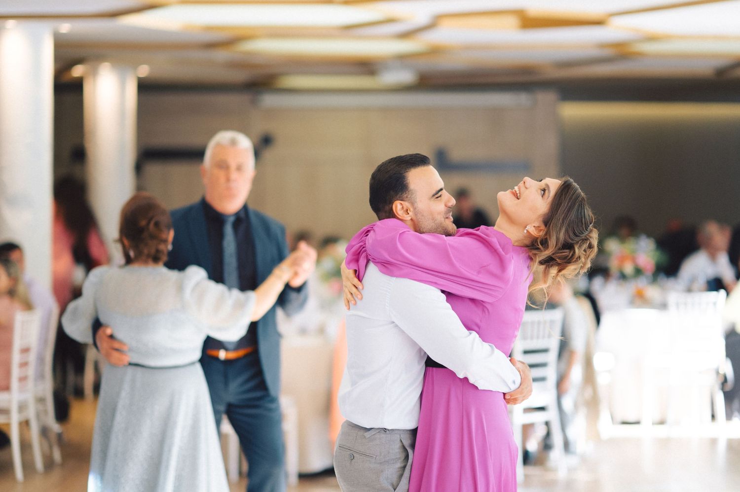 Couples dance together at an indoor venue with people enjoying themselves in the background.