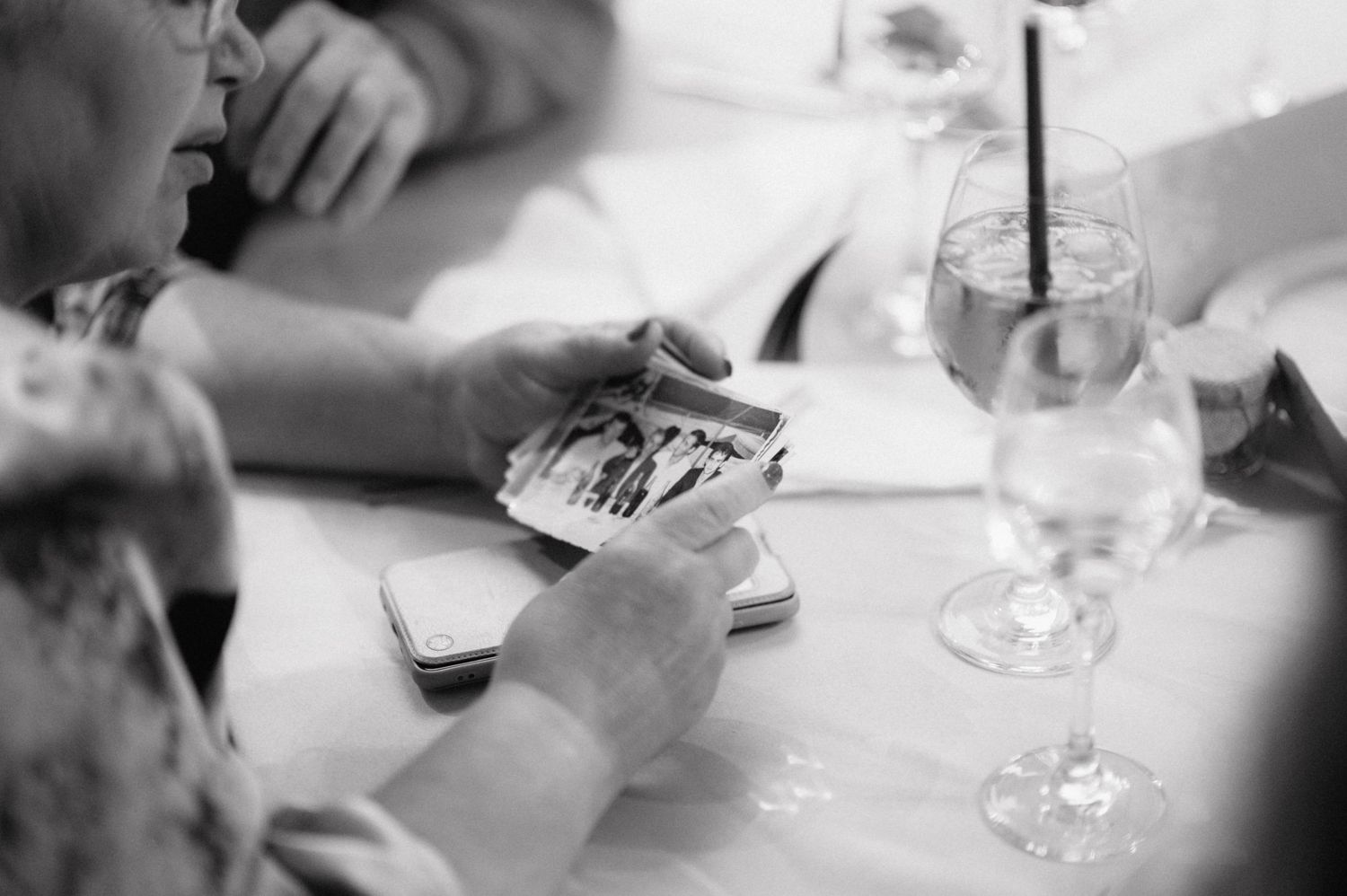 A glass of water sits on a table while someone holds playing cards in a black and white photograph.