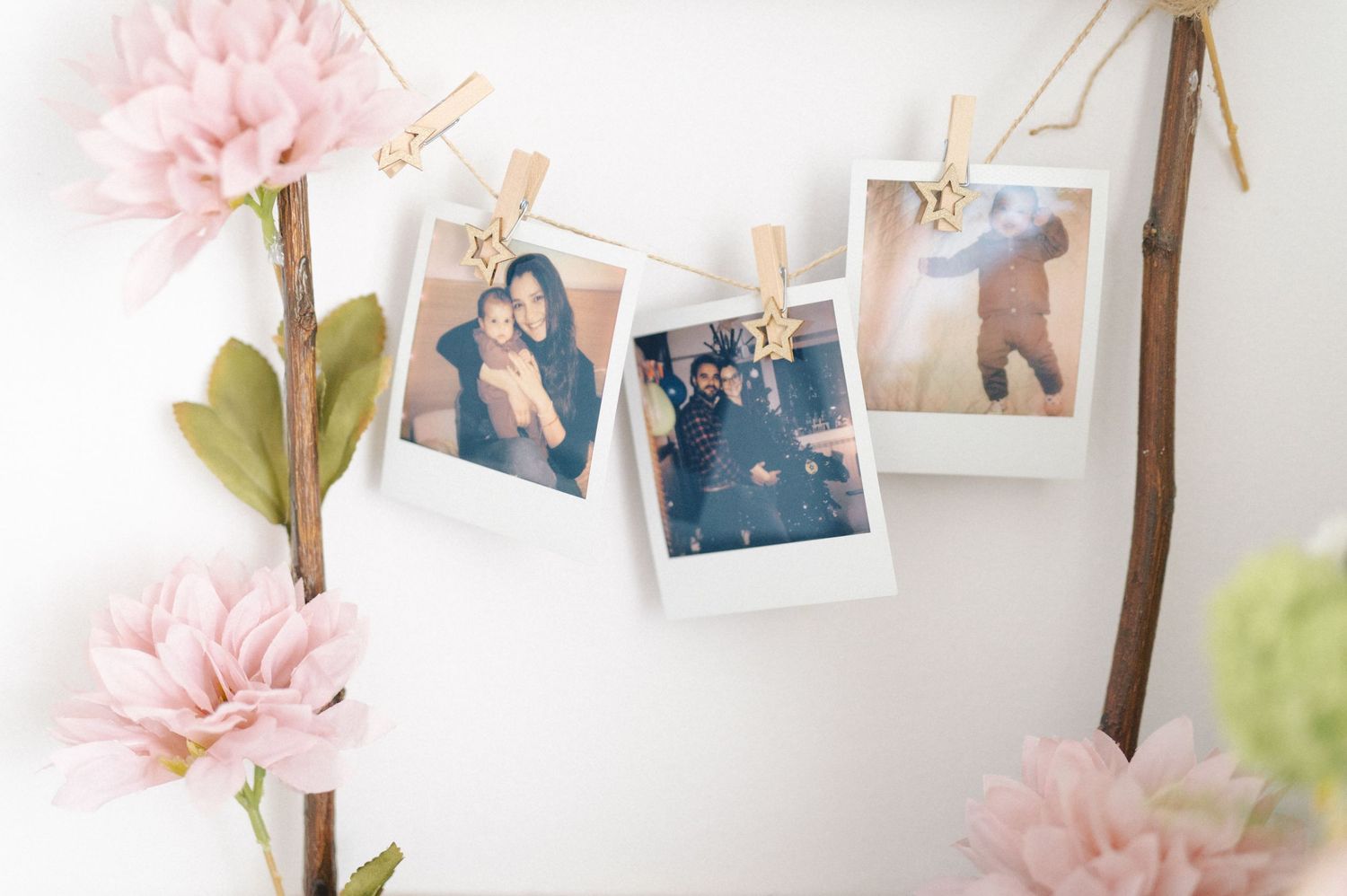 Polaroid photos hang from twine with clothespins alongside pink flowers against a white wall.