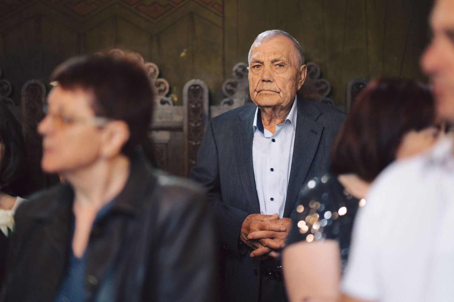An elderly gentleman in a navy suit jacket and white button-down shirt stands thoughtfully among other guests.