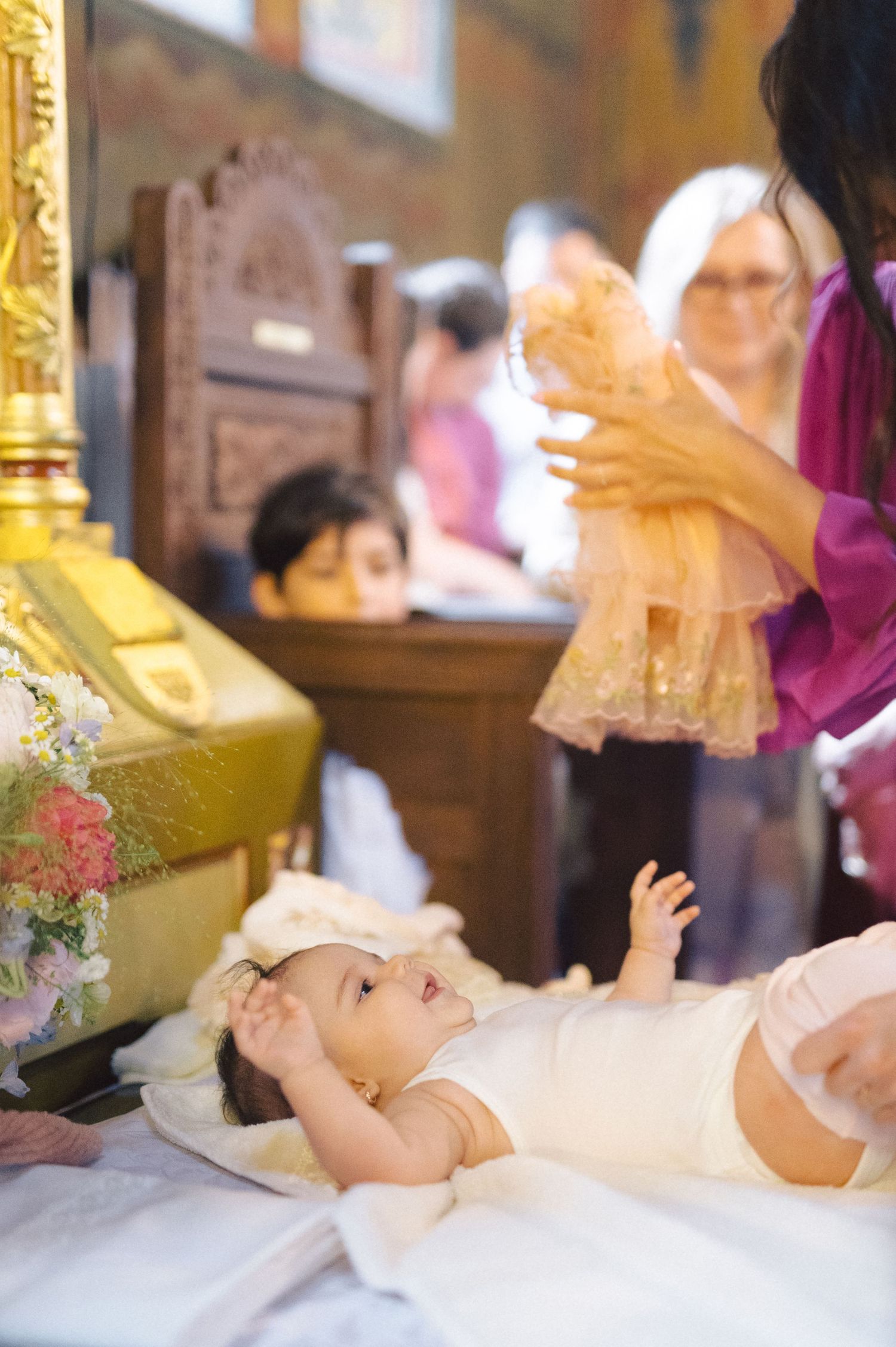 Baby in white dress lies on church altar during christening ceremony as adults gather around.