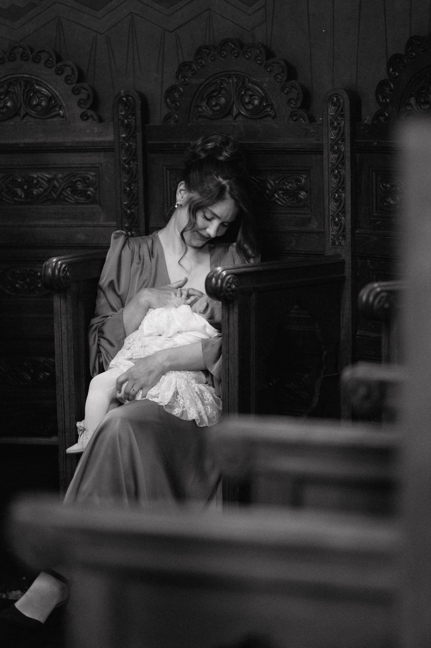 A figure in vintage dress sits in an ornate carved wooden chair holding a baby wrapped in white lace, captured in black and white.
