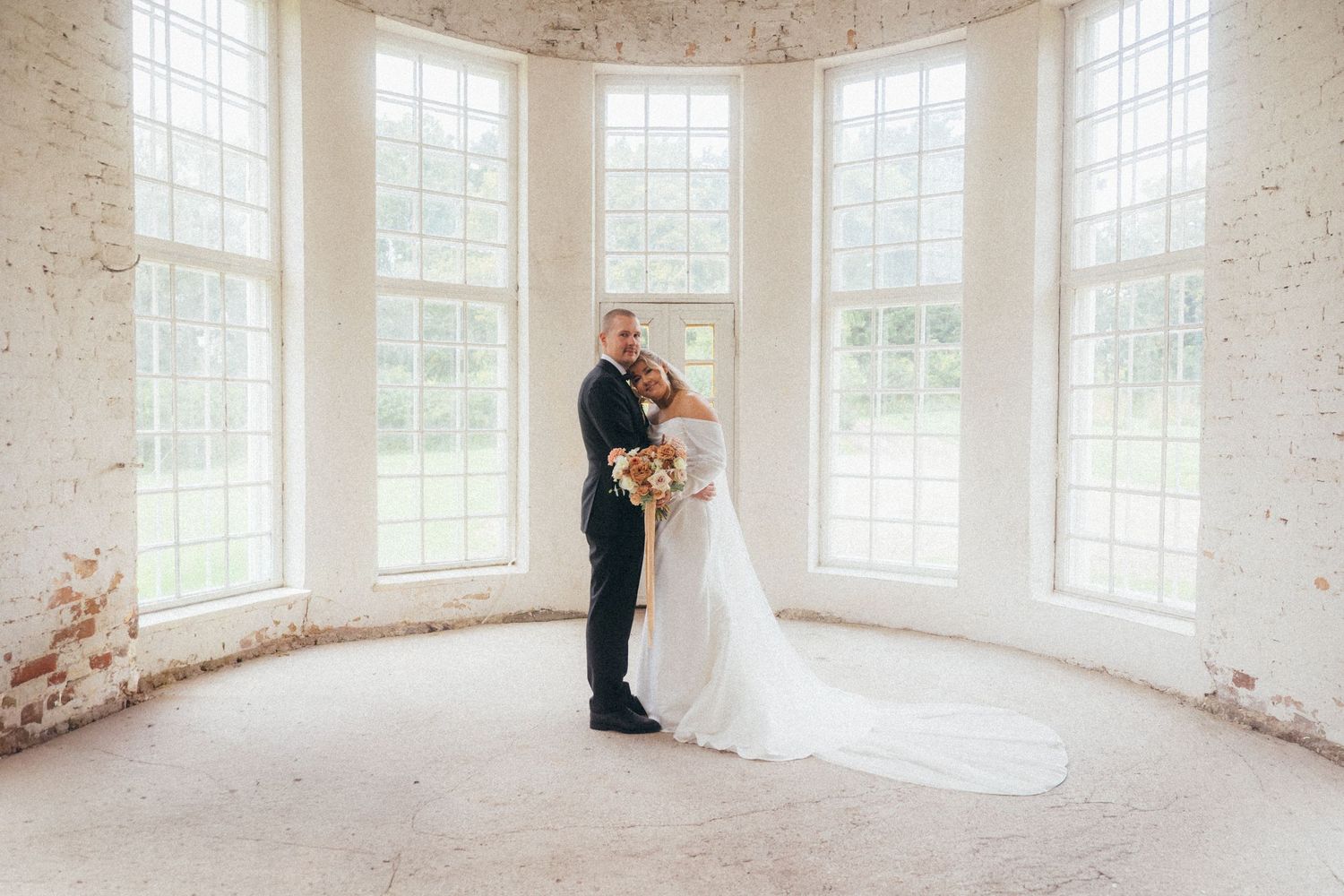 A couple in formal wedding attire shares an intimate moment in a bright sunlit room with large windows.