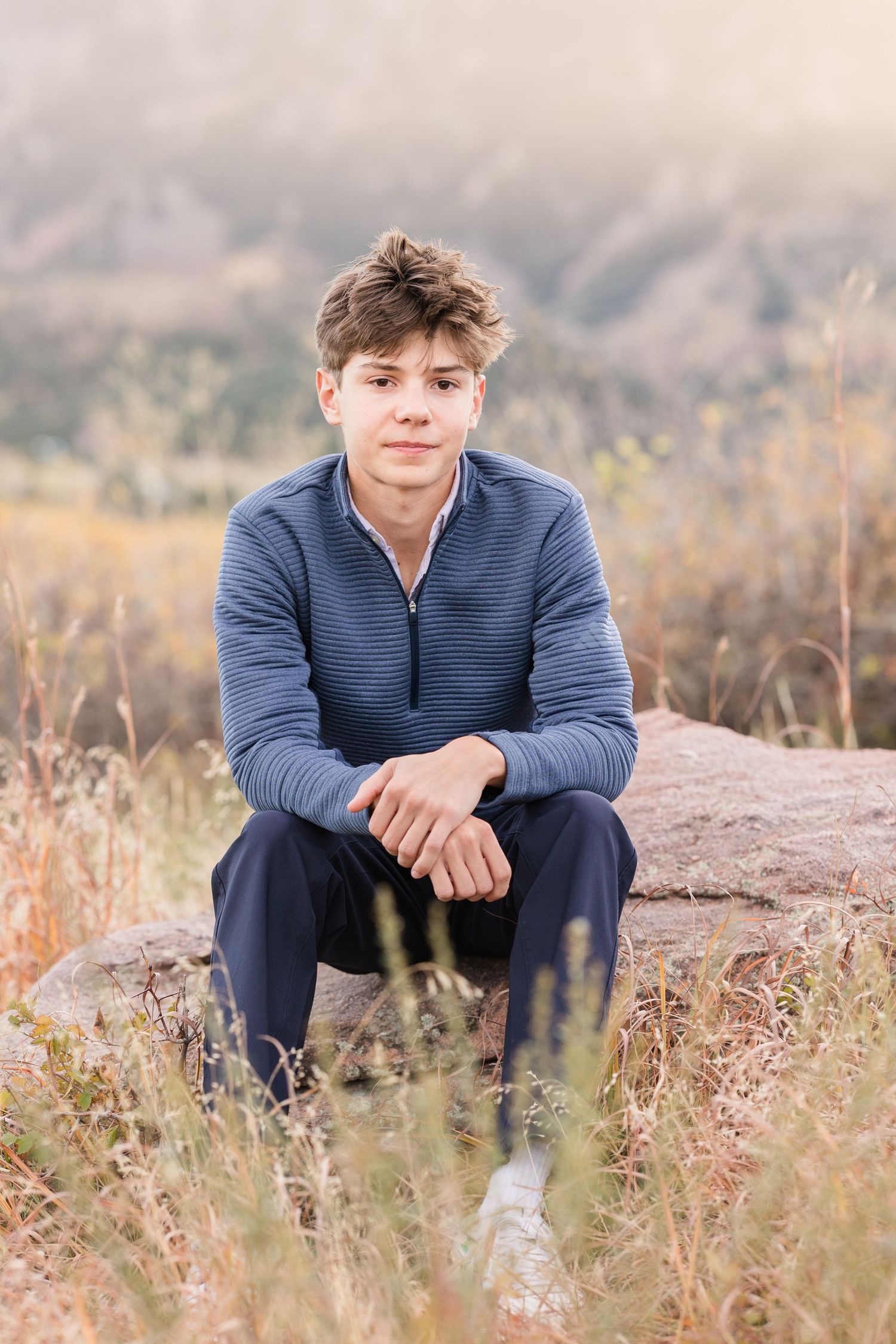 Person in navy sweater sits on rocky outcrop surrounded by tall grass in natural outdoor setting.