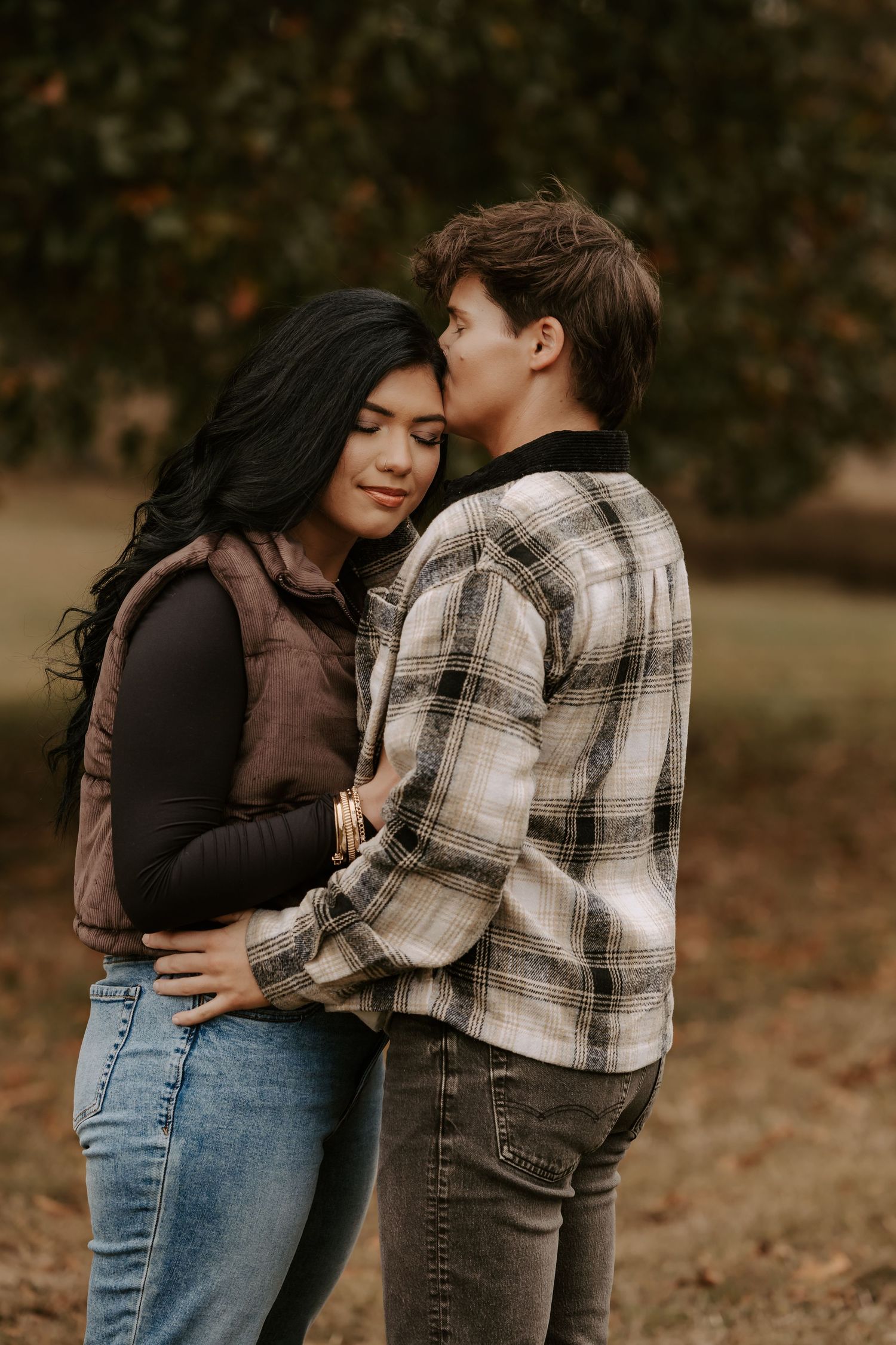 Couple embracing outdoors against a backdrop of autumn trees and fallen leaves.