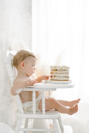Bébé dans une chaise haute antique blanche touchant du doigt son gâteau d’anniversaire pour sa séance fine art smash the cake