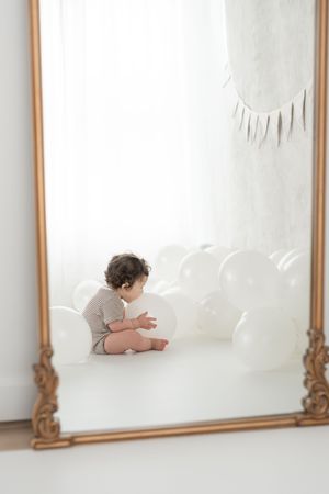 Vue dans un miroir d'un bébé jouant avec des ballons blancs perlés pour sa séance anniversaire fine art au studio de photographie de Québec