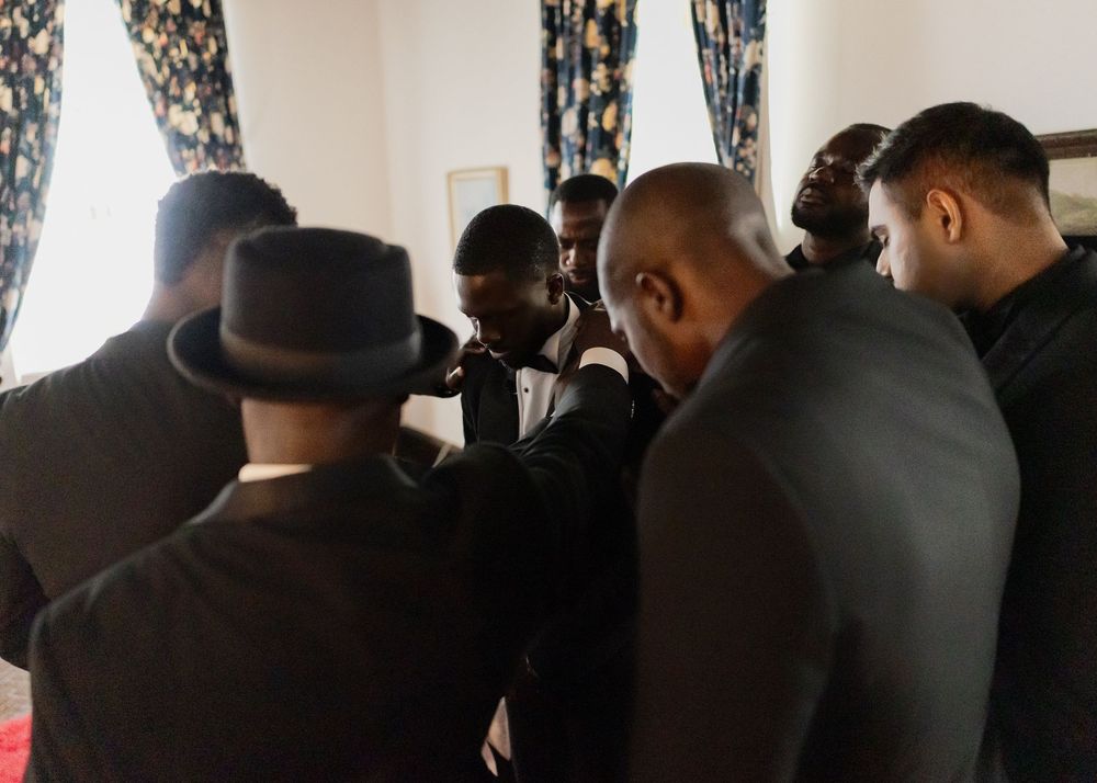 A group of people wearing dark coats and hats stand together in a room with curtained windows.