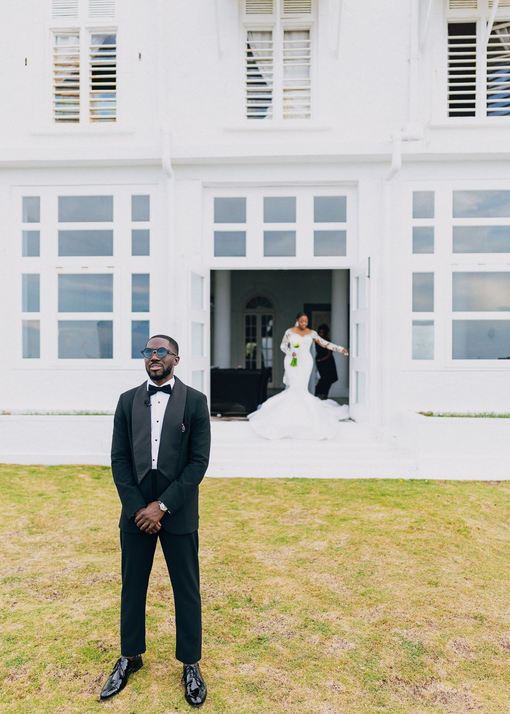 A groom in a classic black tuxedo and bow tie poses on a lawn outside a white building with shuttered windows.