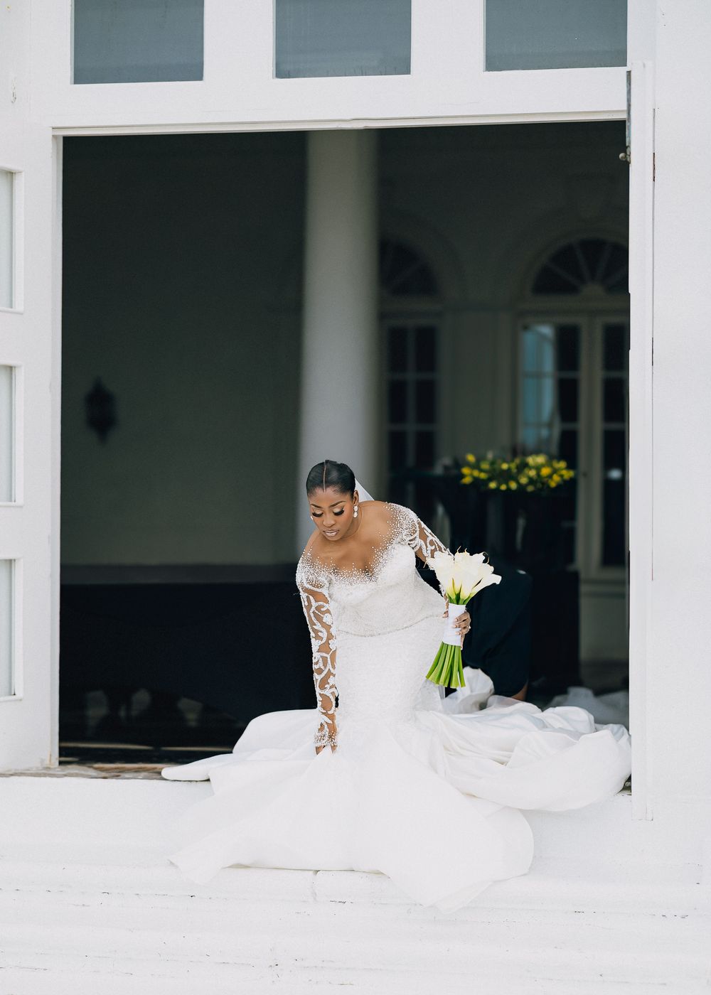 Bride in a flowing white lace gown with long sleeves holds a white bouquet while standing in a bright columned space.