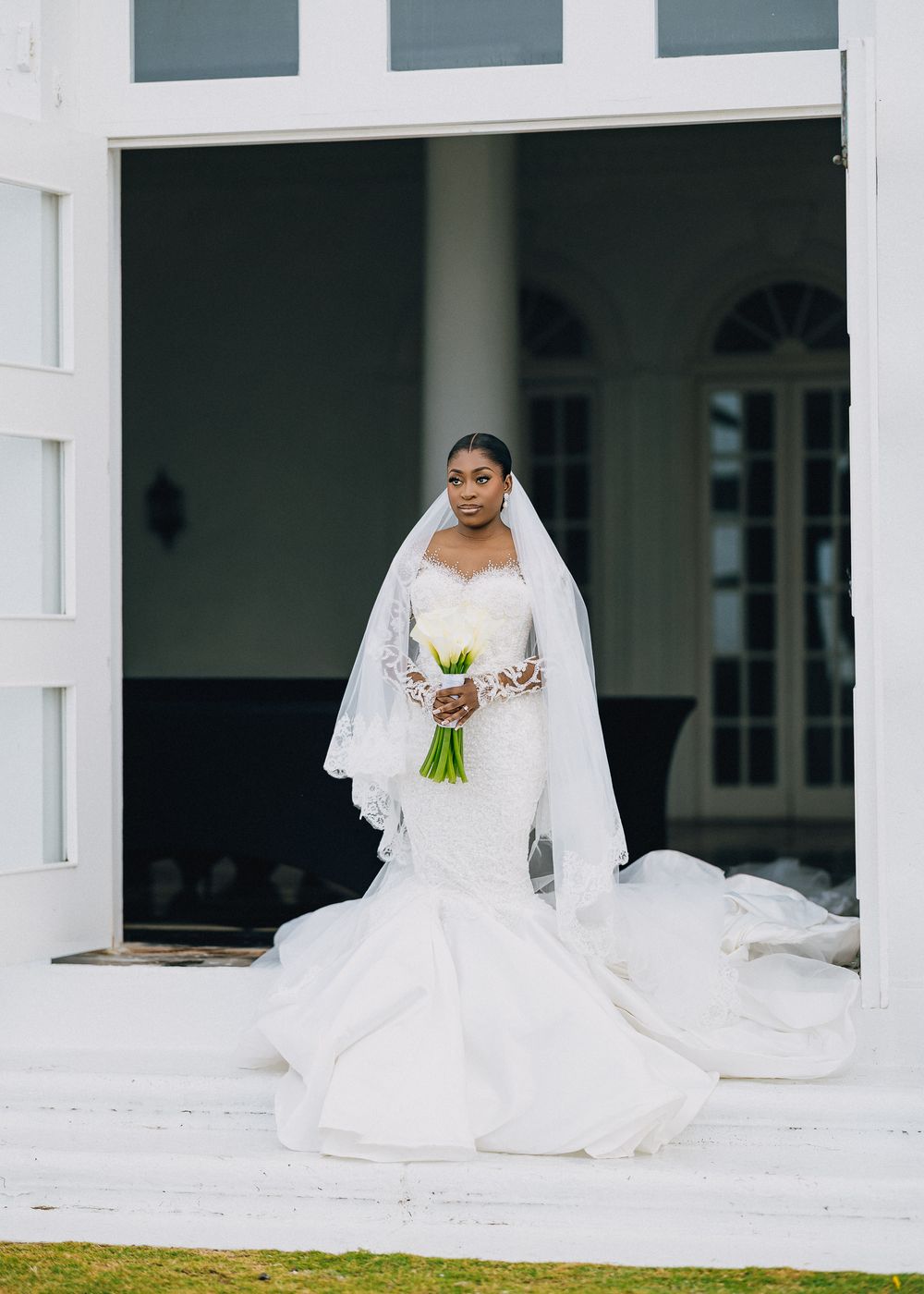 A bride in a flowing white wedding gown sits gracefully on a white porch with elegant columns.