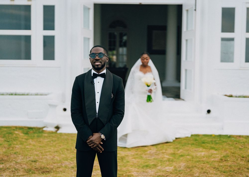 A person in a black tuxedo and bow tie poses outdoors against a white building backdrop.