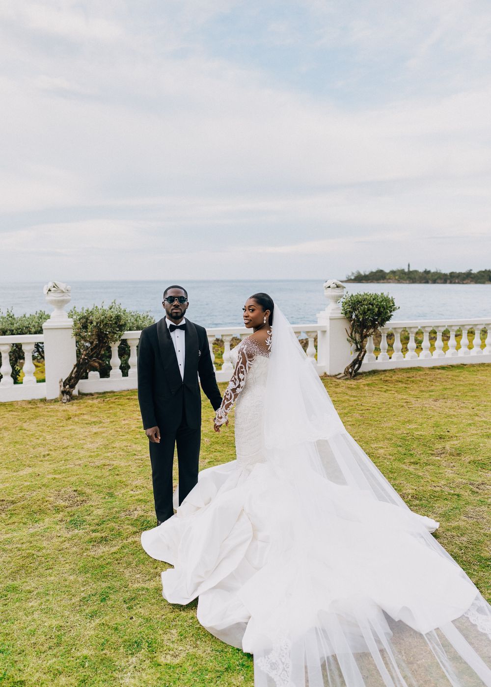 A romantic wedding couple poses on a grassy seaside terrace with white balustrades and ocean views in the background.