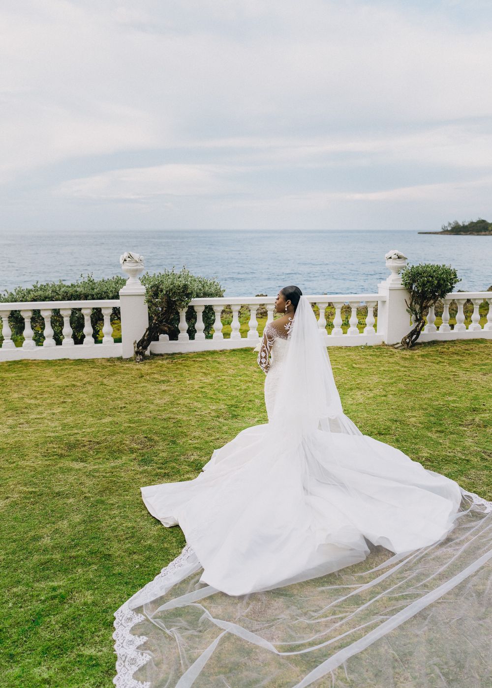 A flowing white wedding gown and veil billow in the breeze on a grassy oceanfront terrace with ornate balustrades.