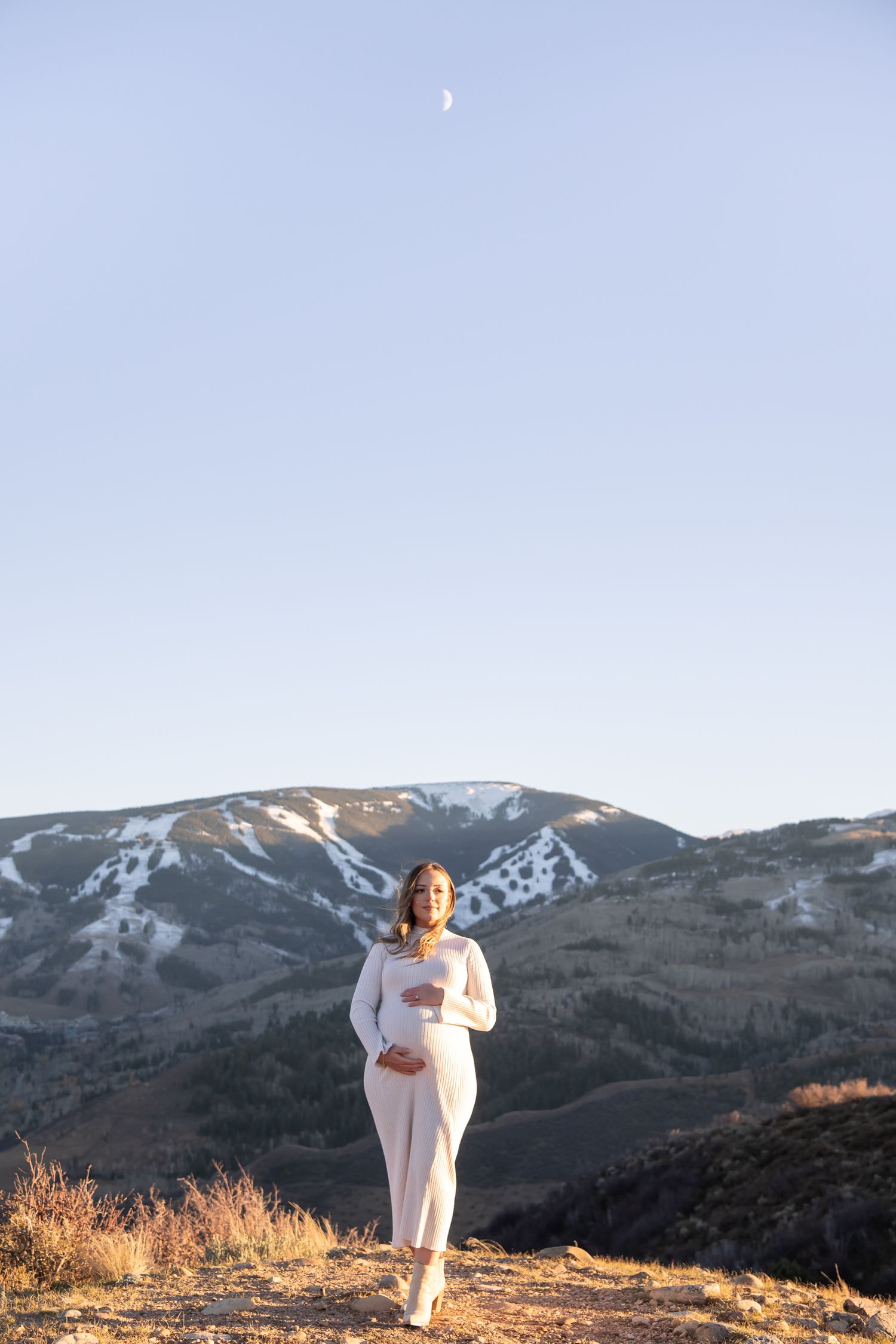 Stunning maternity images with the moon and mountain peaks in the background with a woman dressed in white for Vail maternity photos.