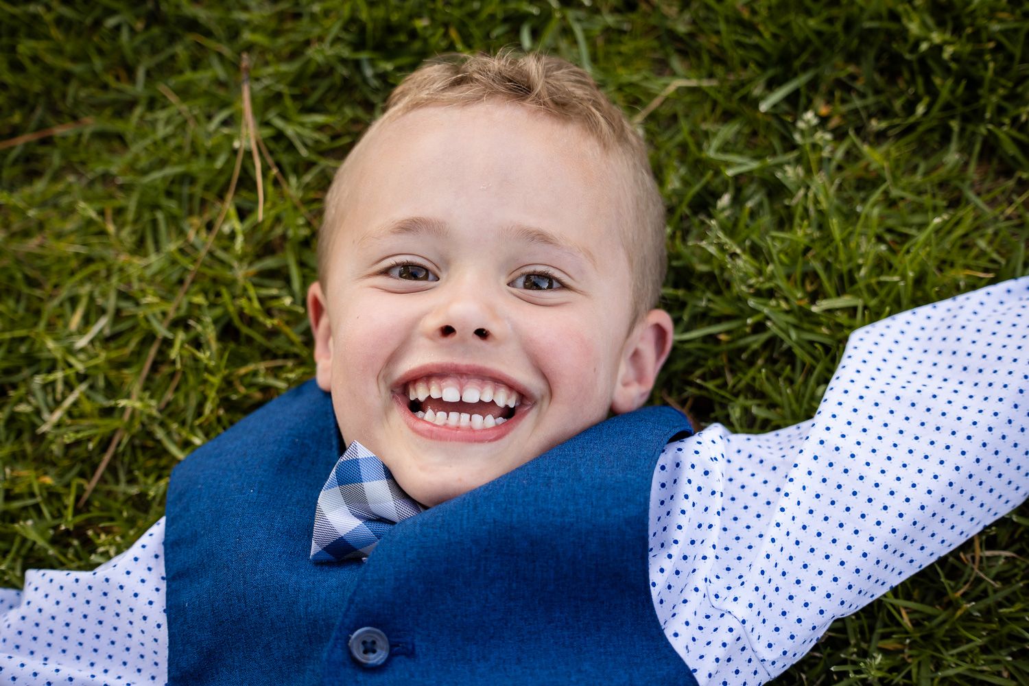 A young boy with a joyful expression lays in green grass wearing blue and white polka dot clothing during his family's photoshoot.