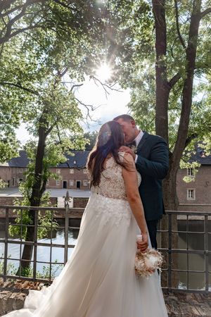 A couple in formal wedding attire shares a romantic kiss under sunlit trees in an outdoor garden setting.